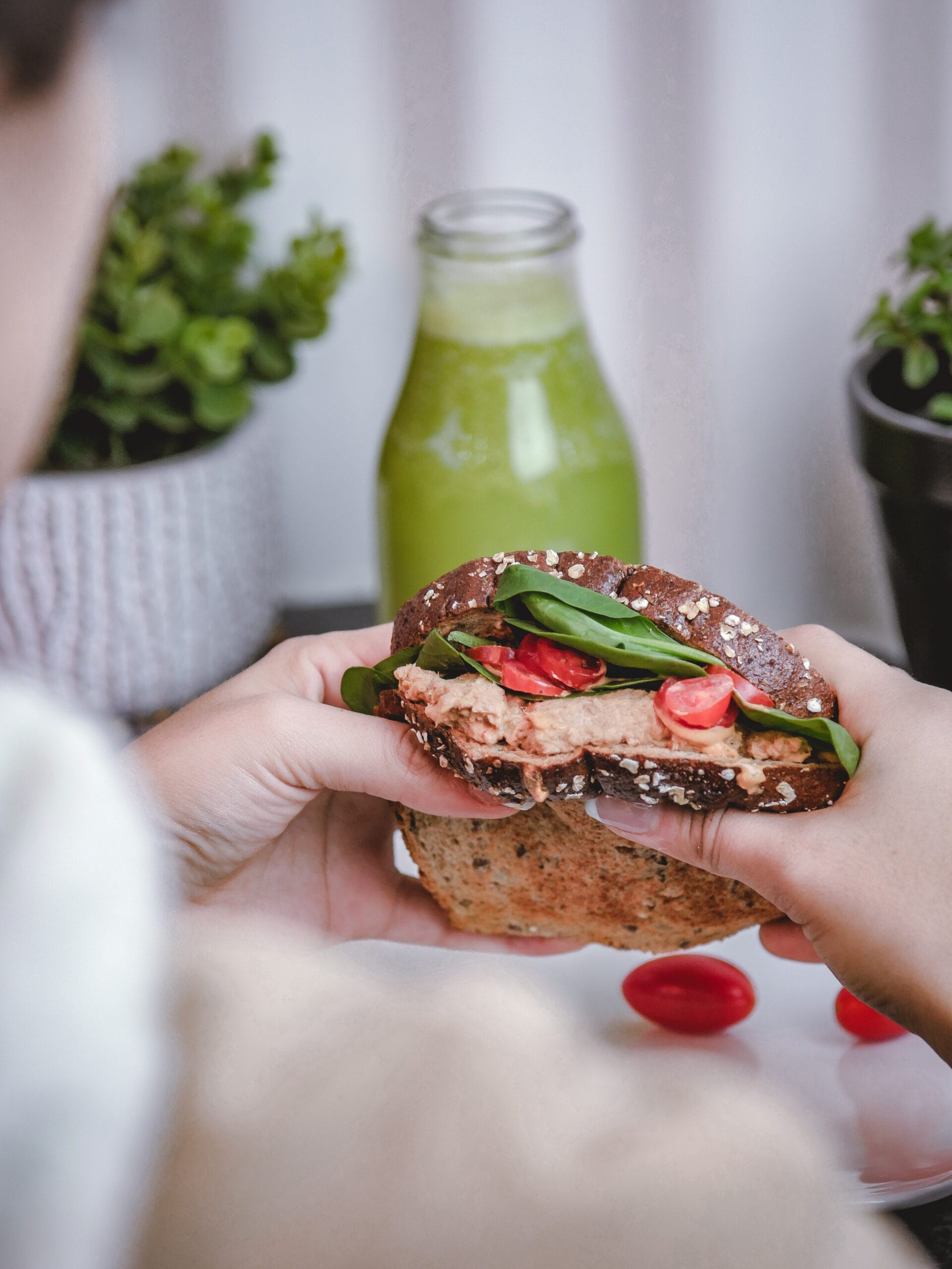 Persoon houdt een volkoren sandwich vast met spread, bladgroenten en tomaat, met groene smoothie op de achtergrond
